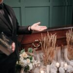 A respectful funeral setting with a closed coffin, flowers, and a man holding a book.