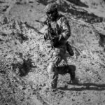 A soldier runs down a sandy hill in full combat gear under harsh desert sun.