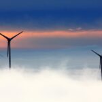 Silhouetted wind turbines rise above clouds against a dramatic sunset sky.