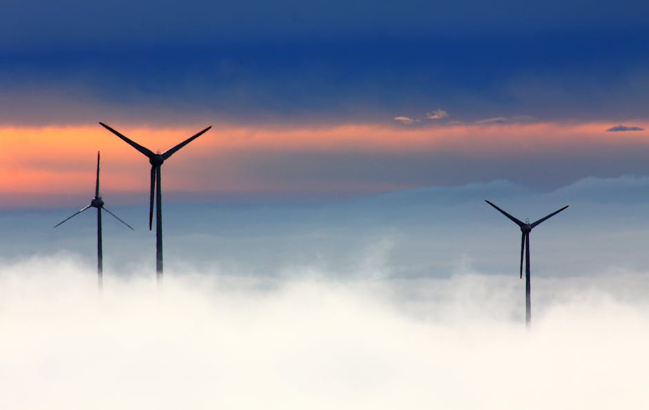 Silhouetted wind turbines rise above clouds against a dramatic sunset sky.