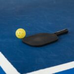 A pickleball paddle and yellow ball on a blue court corner, symbolizing sport and activity.