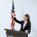 Confident woman in a black suit delivers a speech at a podium with an American flag backdrop.
