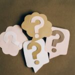 Four paper cutouts of question marks in beige and white on a brown surface, symbolizing inquiry and curiosity.
