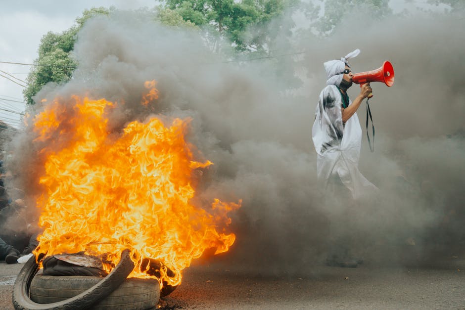 Protest scene with flames and megaphone speaker demanding attention amidst smoke.