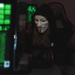 A mysterious hacker wearing a Guy Fawkes mask and black hoodie in a dimly lit room focused on computer screens.