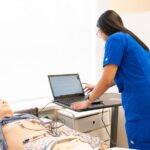 Nurse in training using a laptop with a medical dummy in a clinical simulation lab.
