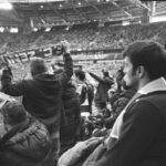 Black and white of group of people in warm clothing standing at stadium and watching match