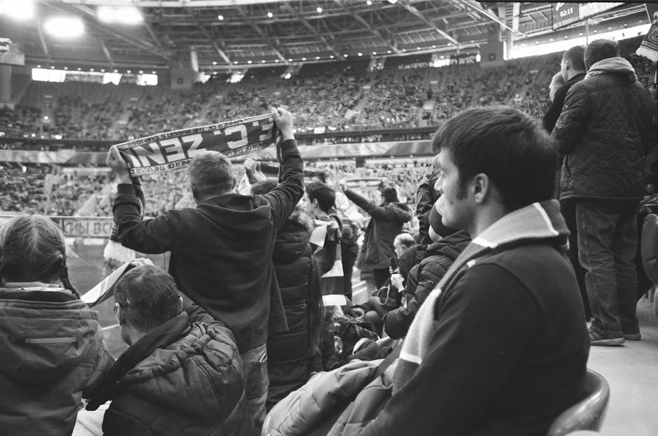 Black and white of group of people in warm clothing standing at stadium and watching match