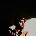 A bearded man in a bucket hat stands in shadow with eyes closed, creating an artistic mood.