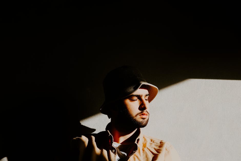 A bearded man in a bucket hat stands in shadow with eyes closed, creating an artistic mood.