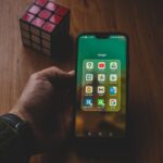 A hand holds a smartphone displaying various apps, next to a Rubik's cube on a wooden surface.