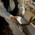 A person reading a book while sitting on a rocky cliff by the sea under natural daylight.