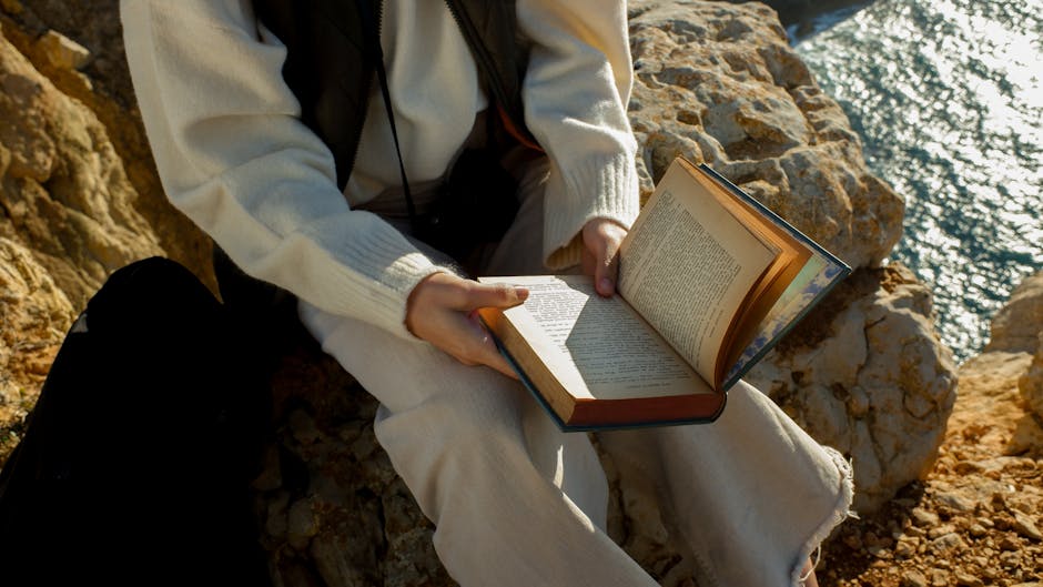 A person reading a book while sitting on a rocky cliff by the sea under natural daylight.