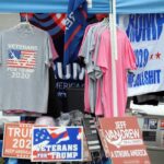 A stall displaying Trump 2020 merchandise including shirts and signs at an outdoor market.