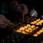 Hands turning takoyaki balls on a hot griddle at a Japanese street food stall.