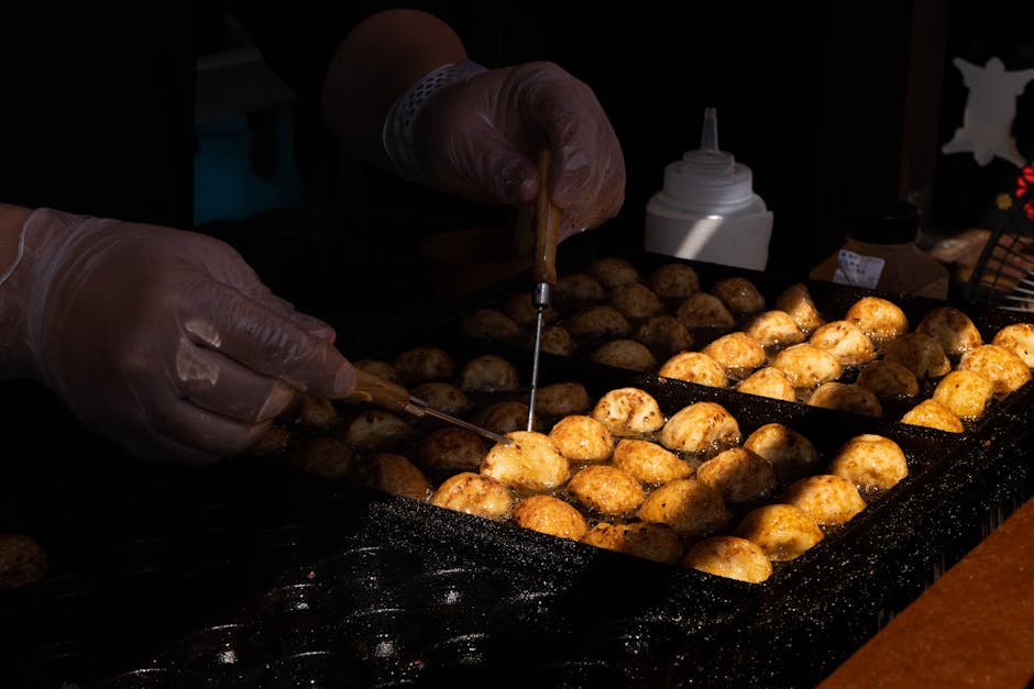 Hands turning takoyaki balls on a hot griddle at a Japanese street food stall.