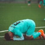 A soccer player kneels in despair on a green artificial turf sports field.