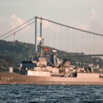 Military vessel with Turkish flags sailing under the Bosphorus Bridge in daylight.