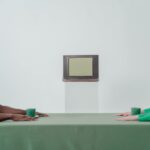 Two people in green clothing sit at a table staring at a vintage CRT TV, creating a minimalist conceptual scene.