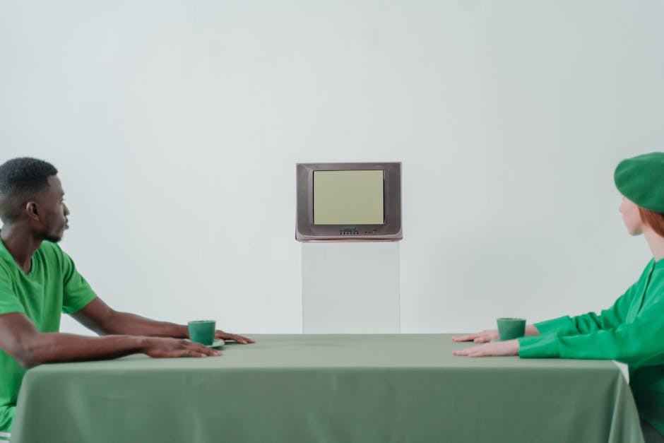 Two people in green clothing sit at a table staring at a vintage CRT TV, creating a minimalist conceptual scene.