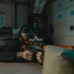 A determined man wearing ear and eye protection aims a rifle at a target in an indoor shooting range.