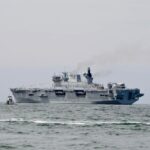 Military aircraft carrier sailing on ocean with visible smoke.