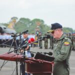 A military officer delivers a speech at an outdoor ceremony with an aircraft and soldiers in the background.