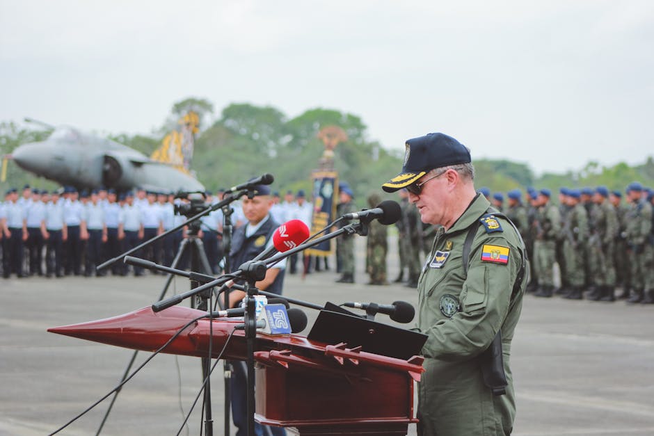 A military officer delivers a speech at an outdoor ceremony with an aircraft and soldiers in the background.