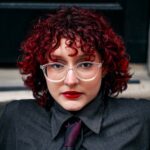 Portrait of a person with red curly hair, wearing glasses and a dark shirt, showcasing a unique style.
