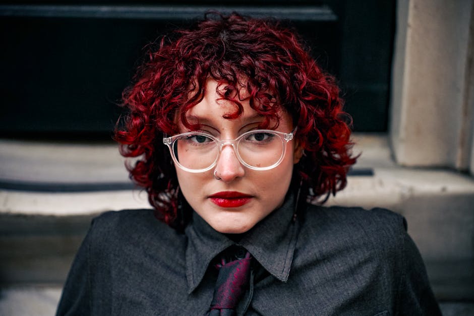 Portrait of a person with red curly hair, wearing glasses and a dark shirt, showcasing a unique style.