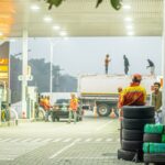 Busy gas station with workers in uniform at twilight, featuring parked cars and a tanker truck.