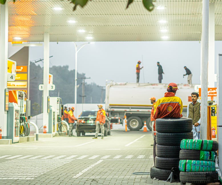 Busy gas station with workers in uniform at twilight, featuring parked cars and a tanker truck.