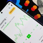 A smartphone showing an investment app with green growth indicators, surrounded by credit cards, US dollars, and a passport.