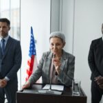 A female politician delivers a speech with bodyguards and an American flag in the background.