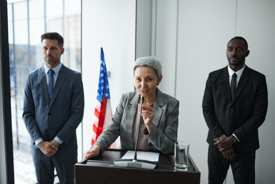 A female politician delivers a speech with bodyguards and an American flag in the background.