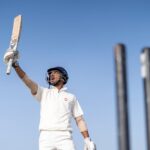 A cricket player exults in victory, raising his bat skyward amidst clear skies.