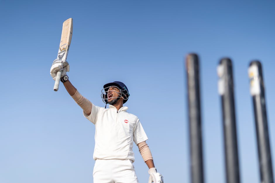 A cricket player exults in victory, raising his bat skyward amidst clear skies.