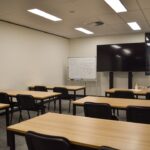 A classroom with tables, chairs, and a large screen.
