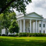 The White House framed by trees and greenery, in Washington, D.C., under a bright sky.