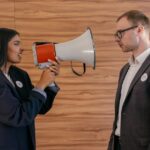 A woman using a megaphone to confront a man in a suit indoors, symbolizing political debate.