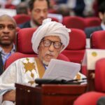 Elderly man in traditional attire reading at a conference with attendees in background.
