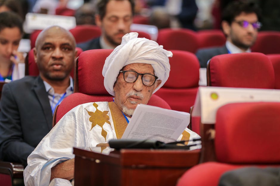 Elderly man in traditional attire reading at a conference with attendees in background.
