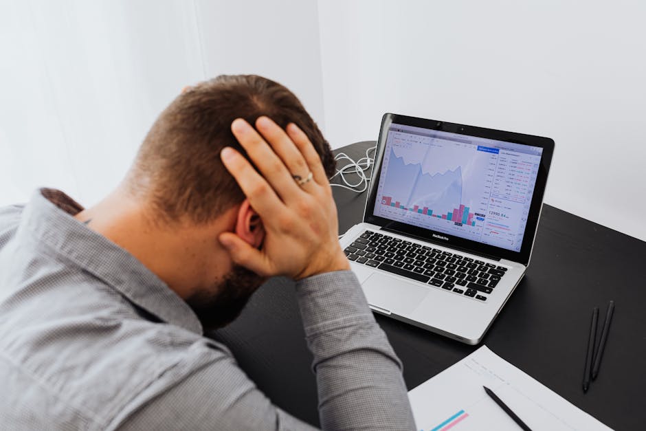Stressed man at desk looking at declining stock charts on laptop, indicating financial loss.