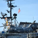 A US Navy helicopter is stationed on the deck of an aircraft carrier with radar equipment in the background.