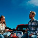 A cheerful couple having a picnic on a truck tailgate under clear blue skies.