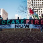 Protesters holding 'Cease Fire Now' banner in urban setting during daytime rally.