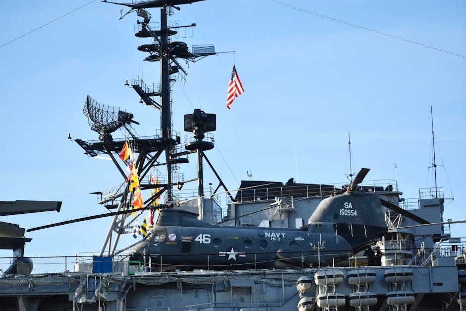 A US Navy helicopter is stationed on the deck of an aircraft carrier with radar equipment in the background.