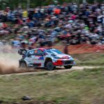 a rally car driving down a dirt road in front of a crowd