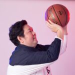 A man in sportswear poses with a basketball against a studio backdrop.