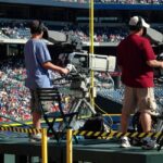 Camera operators capturing a live sports event at a crowded outdoor stadium filled with spectators.